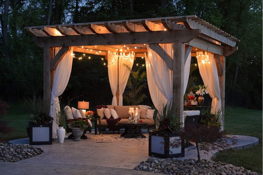 A square gazebo with white curtains and holiday lights in the evening