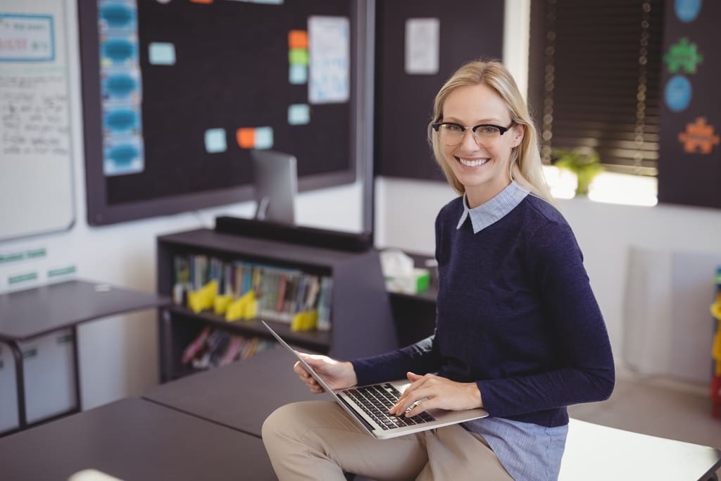 Teacher in classroom on laptop using FutureFund Canva template for penny war fundraiser flyer